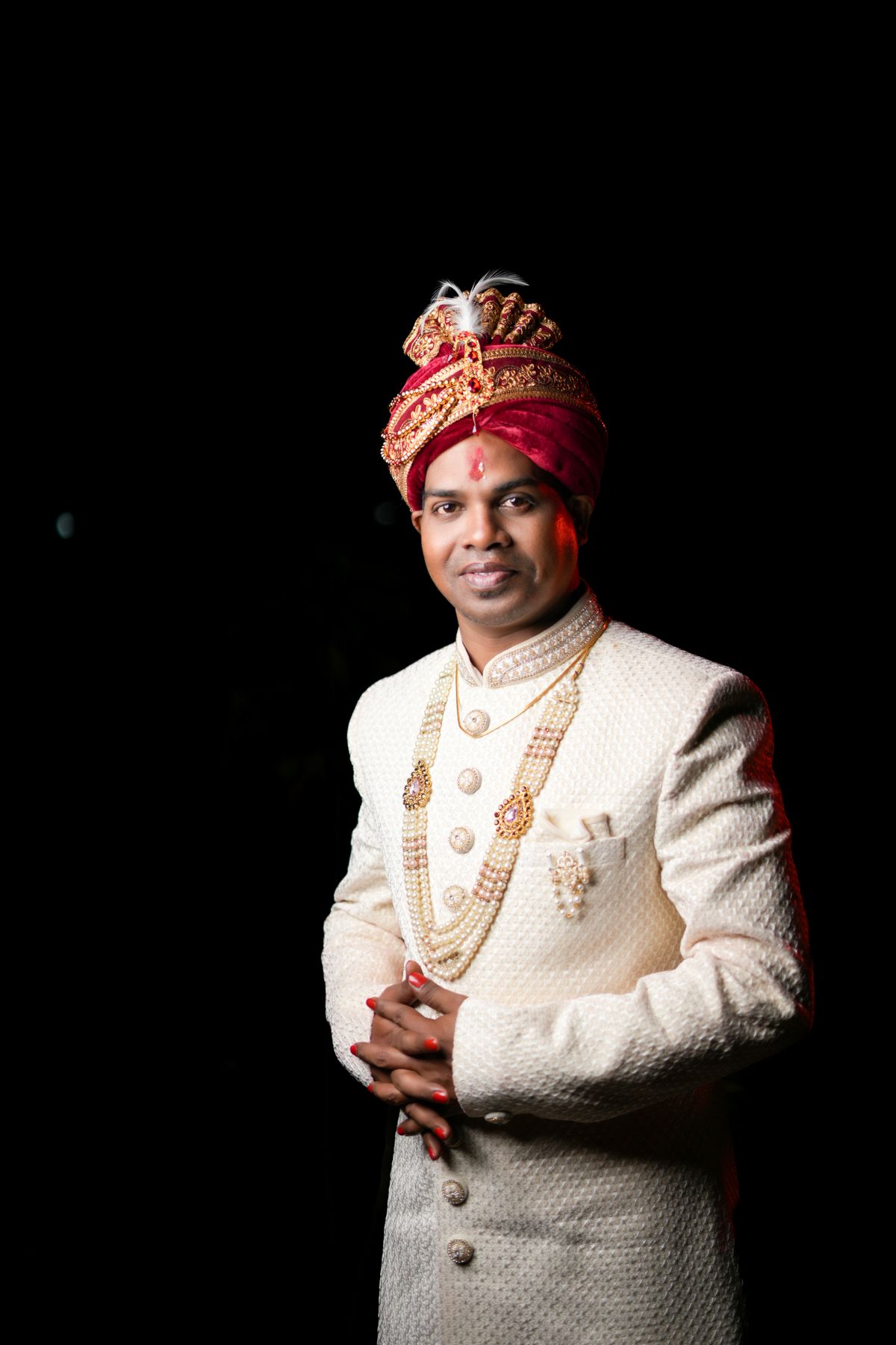 A groom in an ivory sherwani and turban against a dark background