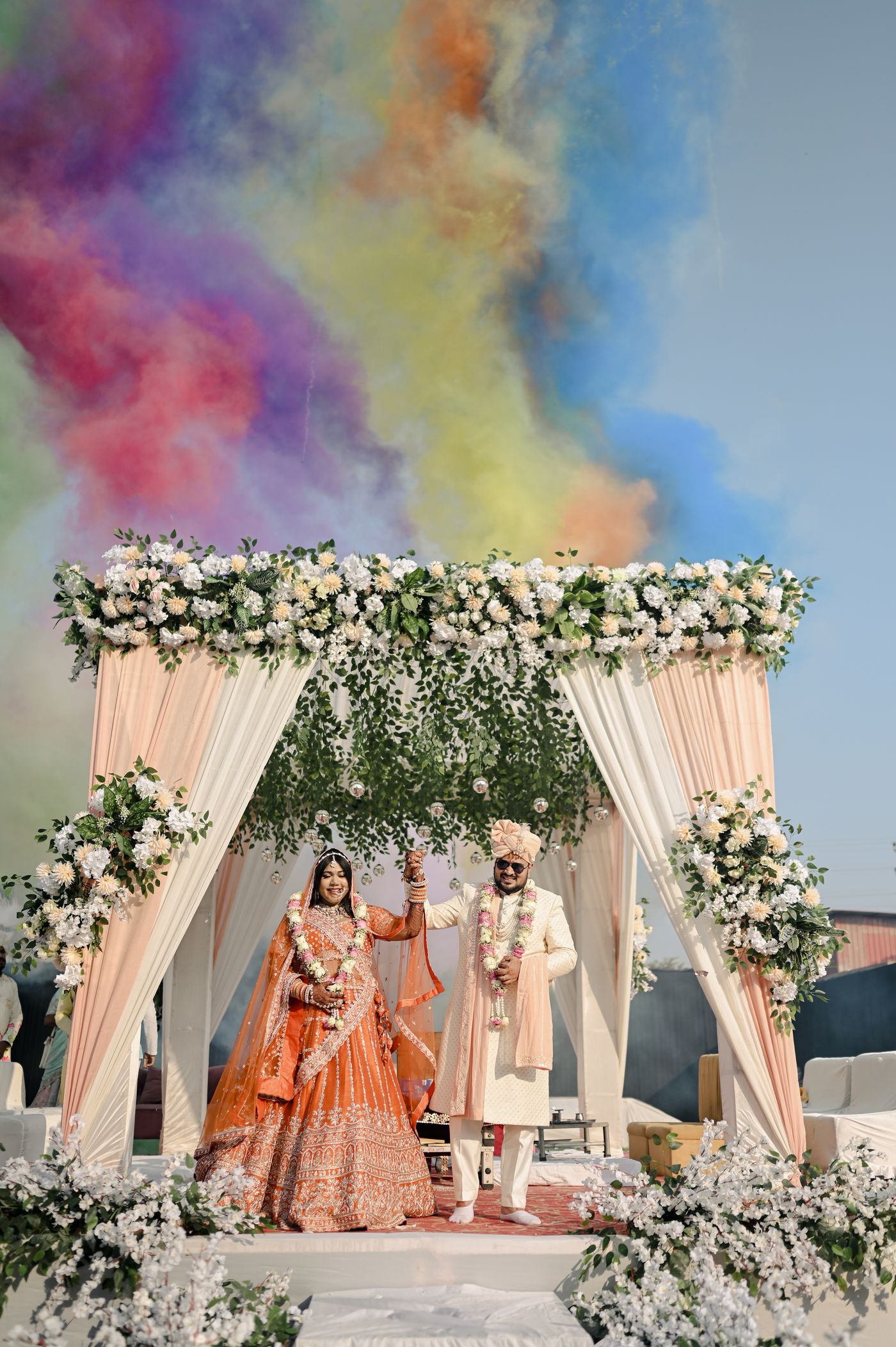 A bride and groom beneath a white floral mandap in a garden